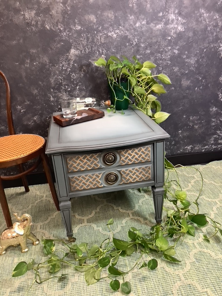 Small gray nightstand with wicker drawers on a textured surface with greenery.