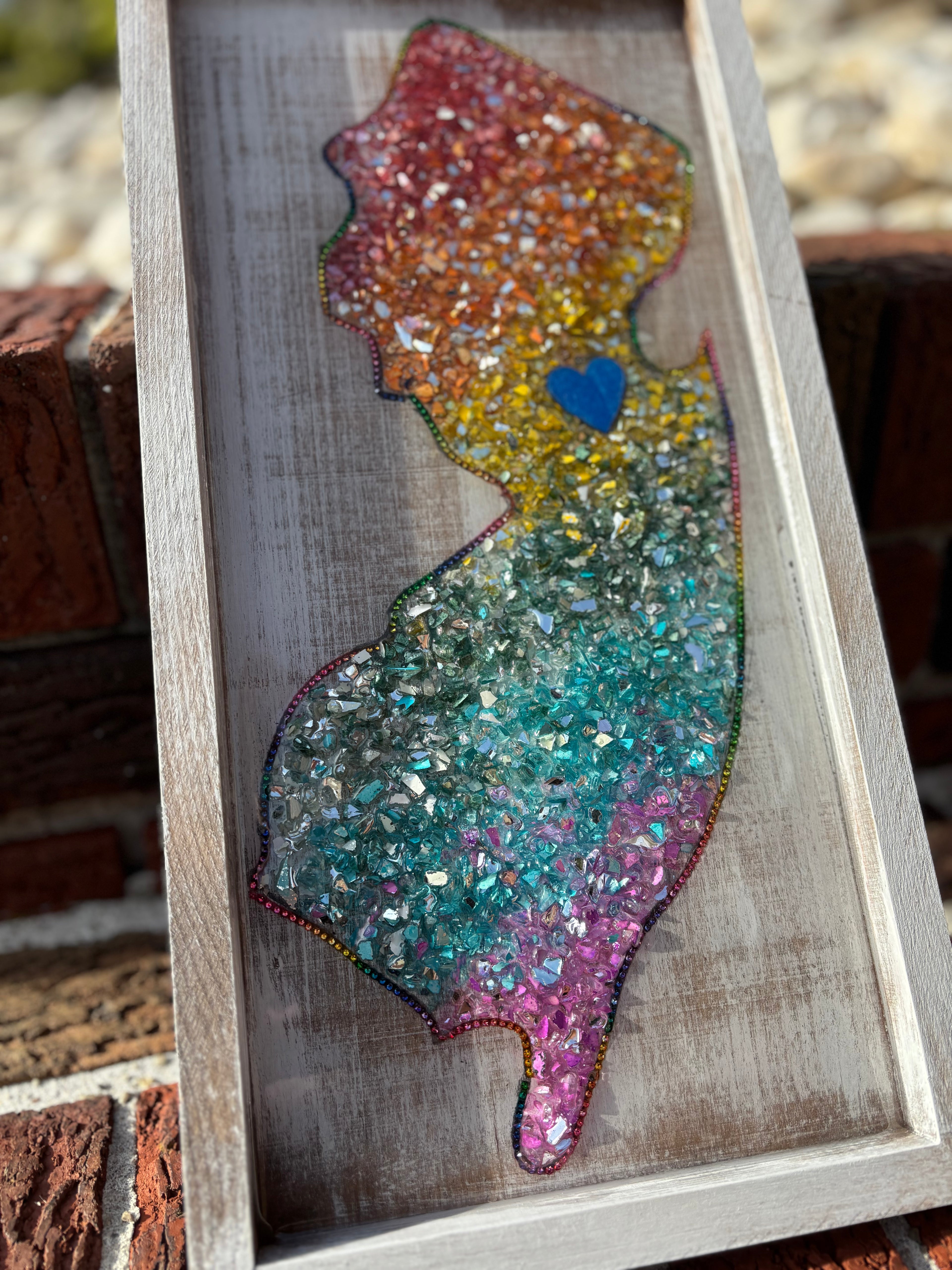 Colorful crushed glass silhouette of a New Jersey with a blue heart on a wooden frame.